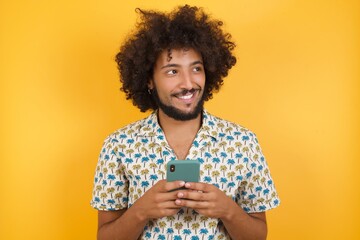 Young man with afro hair over wearing hawaiian shirt standing over yellow background hold telephone...