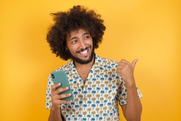 Young man with afro hair over wearing hawaiian shirt standing over yellow background using and texting with smartphone  pointing and showing with thumb up to the side with happy face smiling