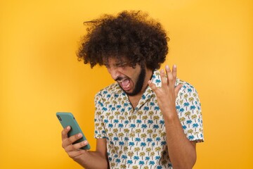 Angry Young man with afro hair over wearing hawaiian shirt standing over yellow background...