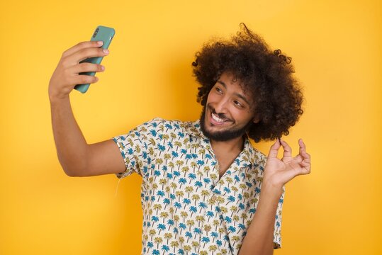 Photo of charming Young man with afro hair over wearing hawaiian shirt standing over yellow background smiling and taking a selfie ready to post it on her social media.