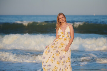 Pretty young blonde woman in dress stands in sunset light with waves behind her