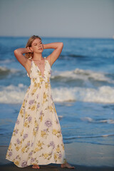 Fototapeta na wymiar Pretty young blonde woman in dress stands in sunset light with waves behind her