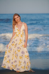 Fototapeta na wymiar Pretty young blonde woman in dress stands in sunset light with waves behind her