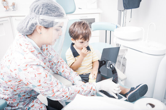 Cute caucasian baby boy sitting on a kids dentist chair interacting with a female dentist in her clinic. Doctor is using protective hair cap and face shield.