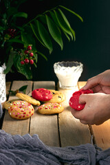 MANO DE HOMBRE PARTIENDO GALLETA ROJA CON MAS GALLETAS EN EL FONDO Y UN VASO DE LECHE SOBRE UNA MESA DE MADERA