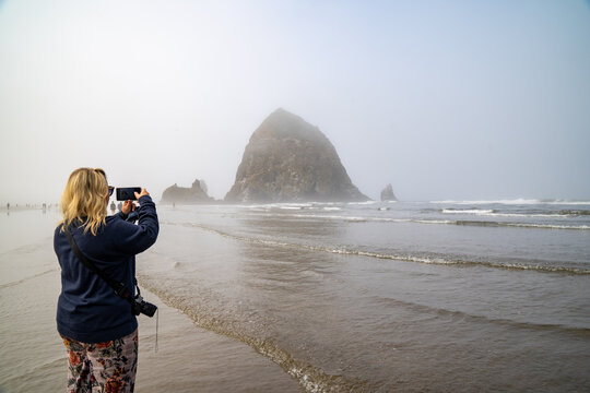 Blonde woman tourists takes photos with her phone of Haystack Rock in Cannon Beach Oregon