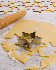 Rolled cookie dough with cookie cutter and rolling pin with baked cookies in the background; Making sugar cookies with dough being cut out and finished cookies