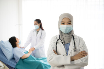 Asian female muslim doctor smiling to camera on background of Asian female doctor taking care of patient in hospital room. Healthcare workers in the Coronavirus Covid19 pandemic