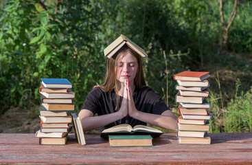 Young girl reading a book and dreams in the garden at a wooden table with a stack of books.