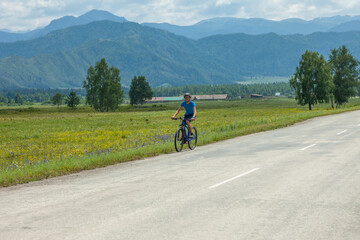 A young cyclist rides a mountain bike along a mountain road past against the backdrop of picturesque blue mountains.