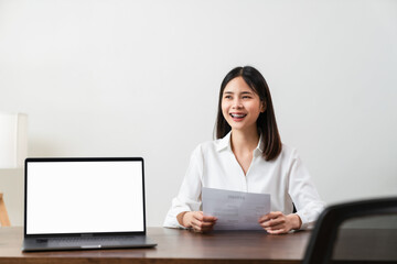 Smiley young Asian woman sitting at a wooden table. Hold resume application information and wait for interviews from company personnel. The digital laptop screen is blank on the desk.