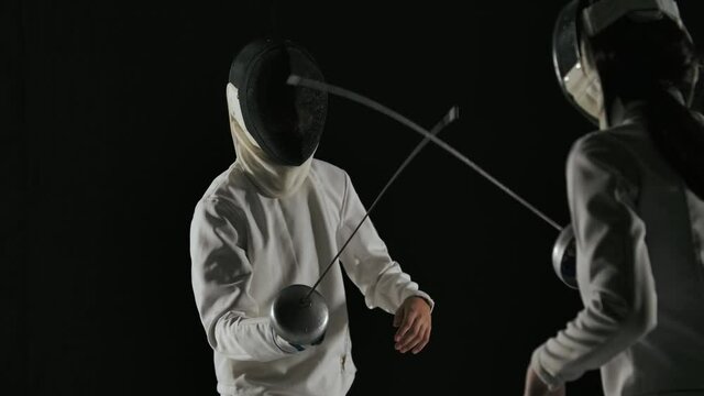 Back view young female fencer in a mask and white suit competes in a duel with an unidentified male fencer. Shot isolated on black background. Slow motion. Close up.