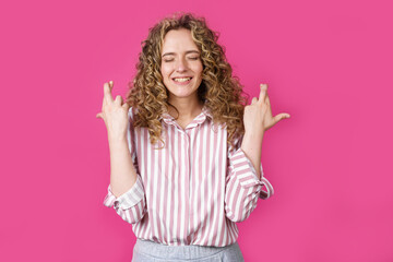 Waiting for a special moment. A portrait of a smiling woman with closed eyes who keeps her fingers crossed. Make a wish. Isolated on a pink background.