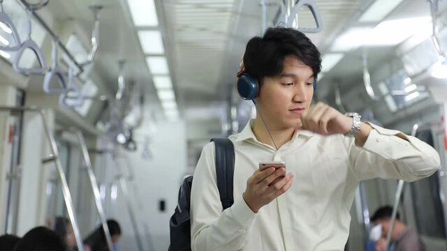 Attractive young Asian man watching video clip from Internet using his mobile phone while keep looking at the time on the watch as he is commuting to work in the morning