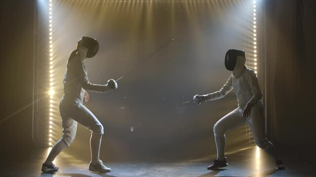 Side view duel of two women fencers. Sportswomen practice strikes, attack and defend. Filmed indoors with studio light. Slow motion.