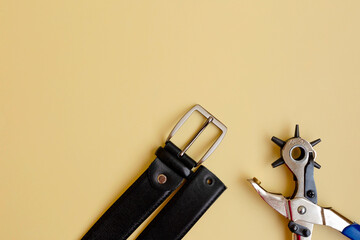 Flat lay of portable leather hole puncher and black belt on the yellow background. Copy space. The...