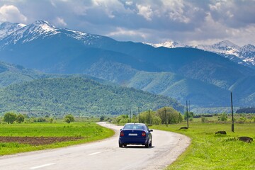 Beautiful view on Carpathian mountains in snow and green forests on the hills in the background and a blue car driving on road in summer in Romania.