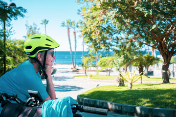 Side view of senior woman with electrice bicycle and helmet resting after excursion in public park with palm trees and horizon over water - healthy lifestyle for retired people