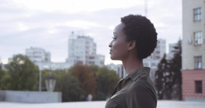 Young attractive international model with stylish hairstyle steps gracefully along street, African foreign student walks in city near buildings, female beautiful face with dark skin, close up portrait