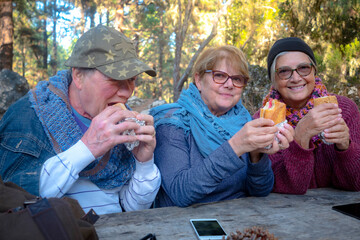 Fototapeta premium Two senior sisters and a husband ienjoying outdoor excursion eating a sandwich at wooden table in pic nic area in the forest. Technological devices on the table