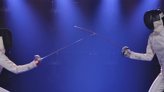 The mastery of the sword of two swordsmen in white suits. Shot in a dark studio against a smoky blue spotlight background. Slow motion. Close up.