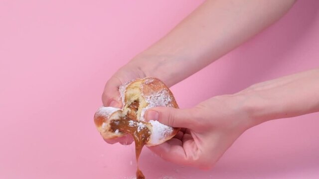Hands tear open a fresh round stuffed donut. Condensed milk is pouring inside. Lots of powdered sugar. Pink background. Delicious food.