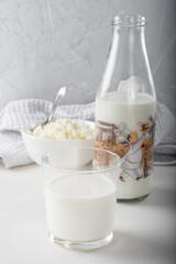 A glass of milk with a bottle of milk and cottage cheese in the background, on a light background.