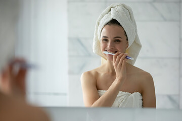 Close up head shot young woman with white bath towel on head cleaning teeth, using toothbrush, standing in bathroom, beautiful female with toothy smile looking in mirror, oral hygiene concept