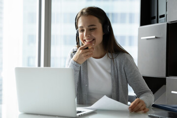 Smiling young woman wearing headset looking at laptop screen, studying or working online, listening...