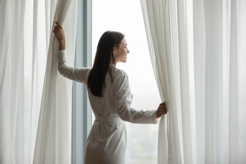Smiling young woman wearing white bathrobe opening curtains in bedroom, standing, beautiful girl looking out large panoramic window, dreaming, visualizing, starting new day, enjoying morning