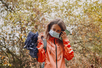 Young caucasian woman putting on a coronavirus mask in the autumn forest. Woman holding smartphone and umbrella in hands.