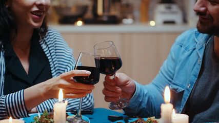 Cheerful couple with red wine glasses sitting at the table in the cozy kitchen. Happy lovers dining together the meal celebrating their anniversary at candle lights
