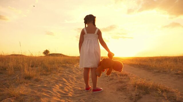 A little girl stands with a teddy bear in her arms in the park. The kid plays with his favorite toy in the rays of the sunset. A lonely child is bored in the park. Dream, lifestyle concept.