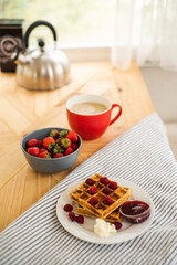 Belgian waffles for breakfast. Waffles with fresh berries and berry jam. Cup of coffee and teapot on the table. Breakfast in the trailer