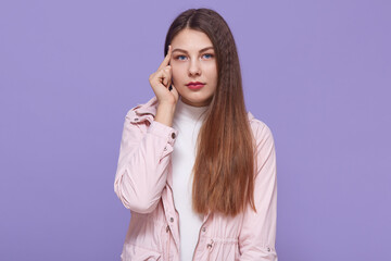Fototapeta premium Young thoughtful female has serious pensive expression, keeping finger near temple, wearing pale pink jacket, stands against lilac background, looks at camera.