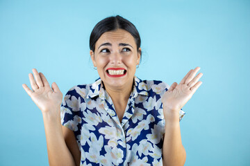 Young beautiful woman over isolated blue background clueless and confused expression with arms and hands raised.