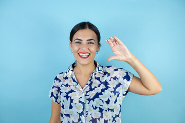 Young beautiful woman standing over blue isolated background smiling and confident gesturing with hand doing small size sign with fingers looking and the camera. Measure concept.