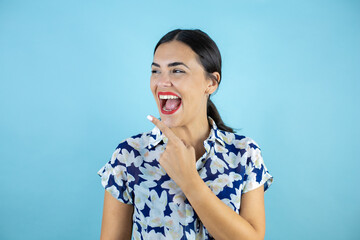 Young beautiful woman standing over isolated blue background smiling looking to the side and pointing to the empty space with one finger.