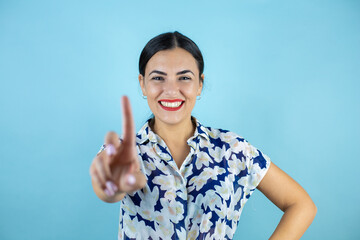 Young beautiful woman over isolated blue background showing and pointing up with finger number one while smiling confident and happy.