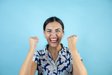 Young beautiful woman standing over isolated blue background screaming with her fists in the air with a gesture of victory.