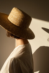 Young pretty woman in straw hat and white dress / sundress against the wall. Silhouette in sunlight. Shadows on the wall. Minimal fashion design concept.