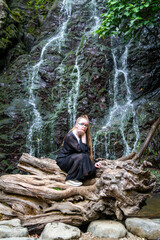 Young girl in black posing squatting on a large dry tree against the background of a mountain waterfall