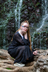 Young girl in black posing squatting on a large dry tree against the background of a mountain waterfall