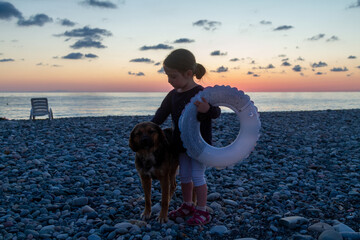Little girl with a lifebuoy stroking a dog on a pebble beach of the Black Sea against a sunset background