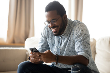 Smiling African American man using phone, sitting on couch, happy young male holding smartphone,...