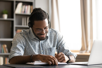 Smiling motivated African American student wearing headphones listening to lecture, writing notes in notebook, sitting at work desk, learning language online, watching webinar, e-learning concept