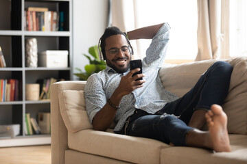Happy African American man wearing headphones using phone, lying on cozy couch at home, smiling...