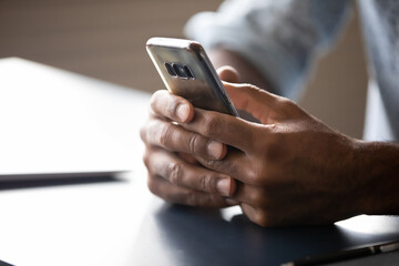 Close up African American man holding phone, browsing mobile device apps, sitting at work desk, male hands touching smartphone, typing on screen, texting in social network, surfing internet