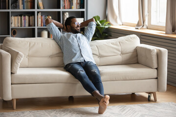 Smiling dreamy African American man sitting on cozy couch with hands behind head, thinking about good future, looking to aside, dreaming about new opportunities, enjoying lazy weekend at home