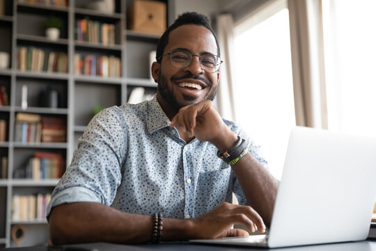 Head shot portrait laughing African American businessman wearing glasses sitting at work desk with laptop, looking at camera, handsome happy young man freelancer blogger working online
